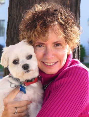 A smiling woman with short, curly hair and a hot pink turtleneck holds a small white dog.