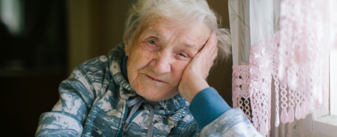 An elderly Holocaust survivor sits at her kitchen table, next to an old window with a lace curtain.