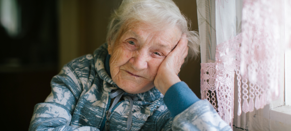 An elderly Holocaust survivor sits at her kitchen table, next to an old window with a lace curtain.